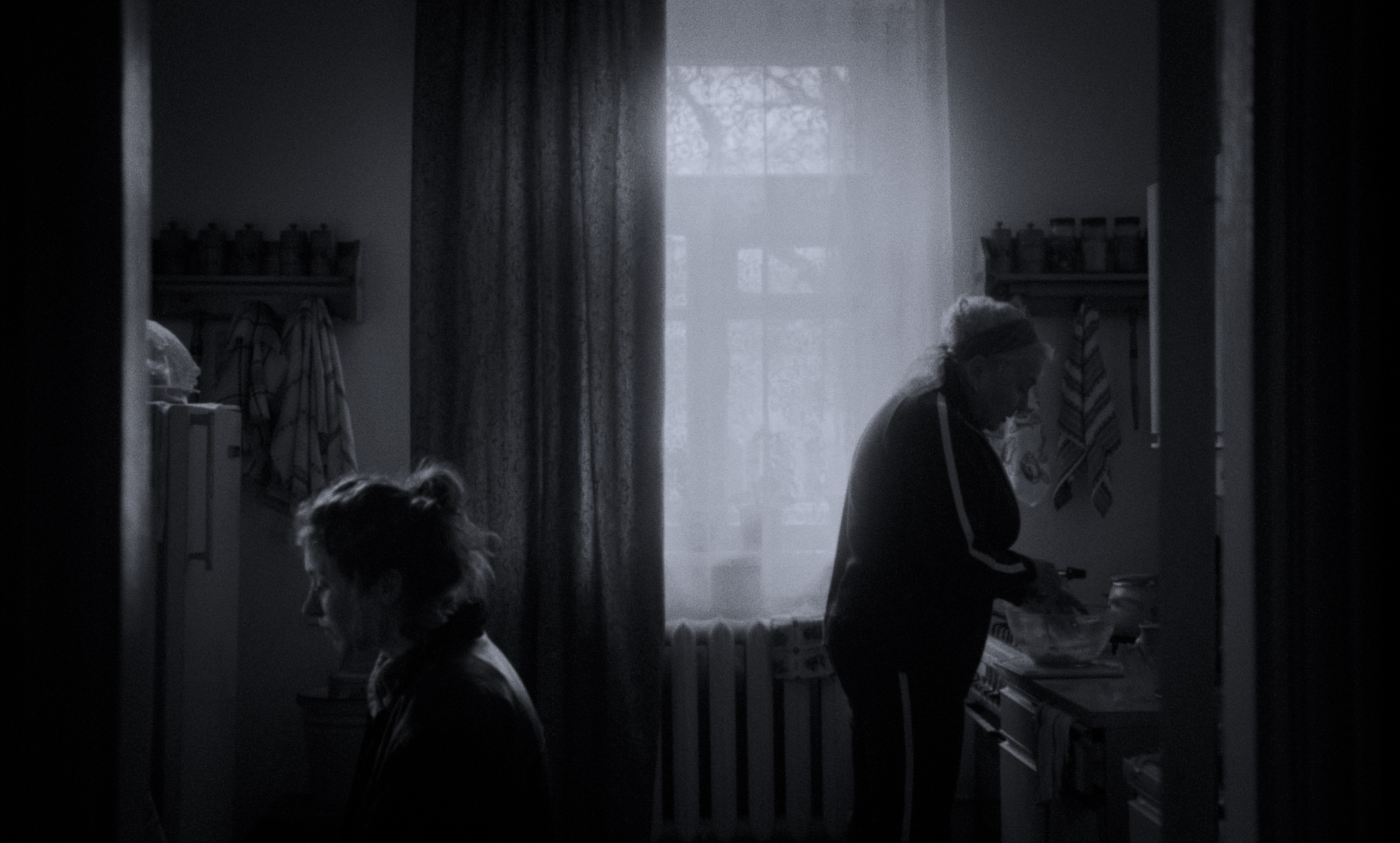 Black-and-white still: two women in a small kitchen, one cooking, one sitting in shadow — Kira’s Dre