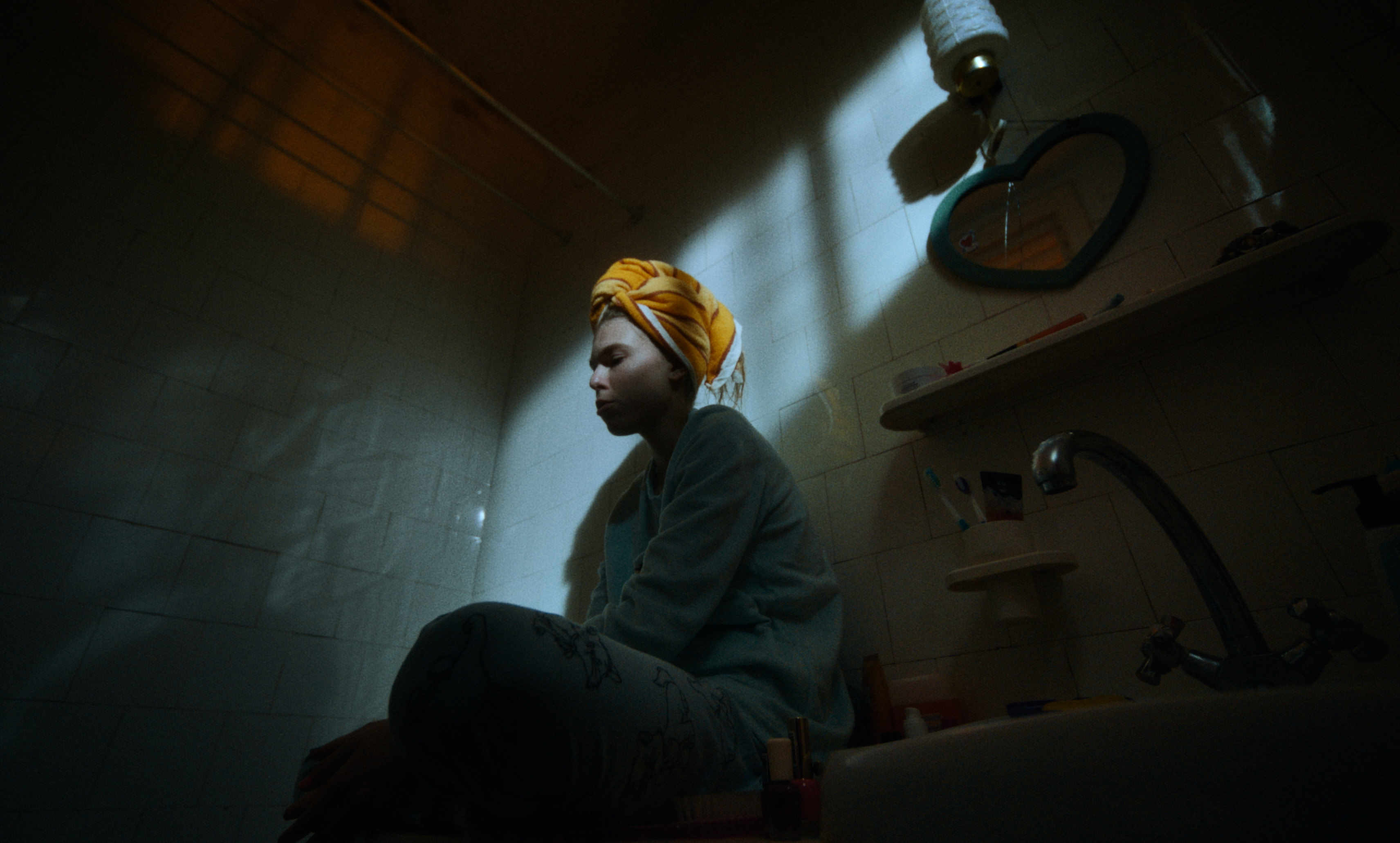 A young woman sits on the edge of a bathtub in dim light, towel wrapped around her head — a still fr