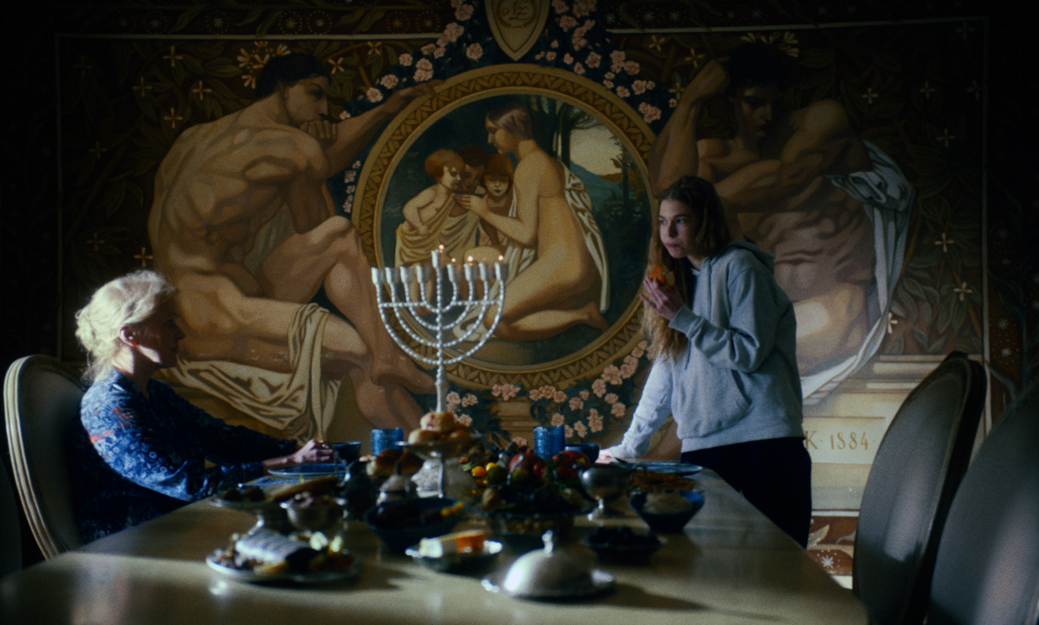 Two women at a dinner table with a lit menorah before a painted mural — a still from Kira’s Dream.