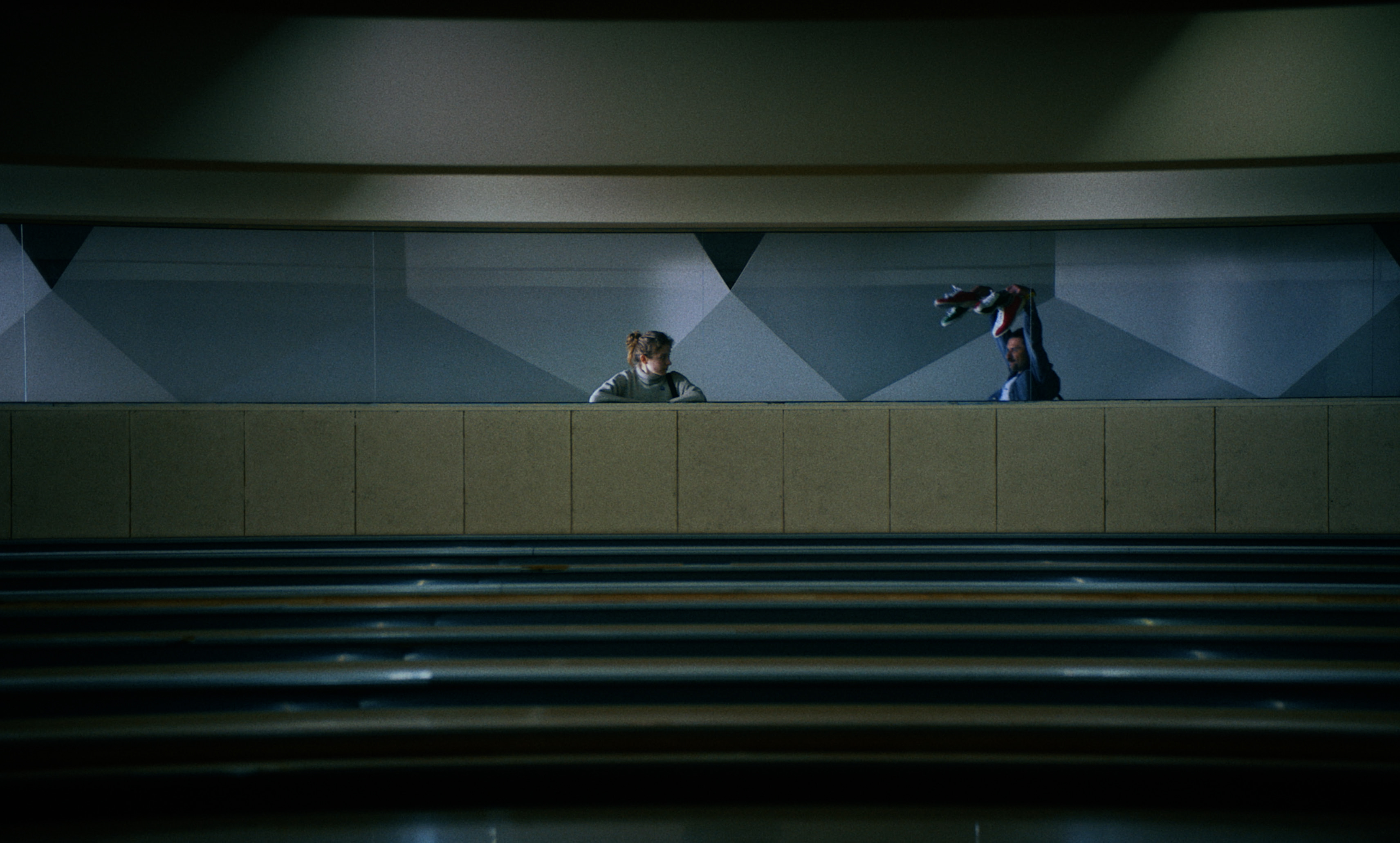 A woman and a man face each other above bowling lanes, framed by geometric walls — a still from Kira