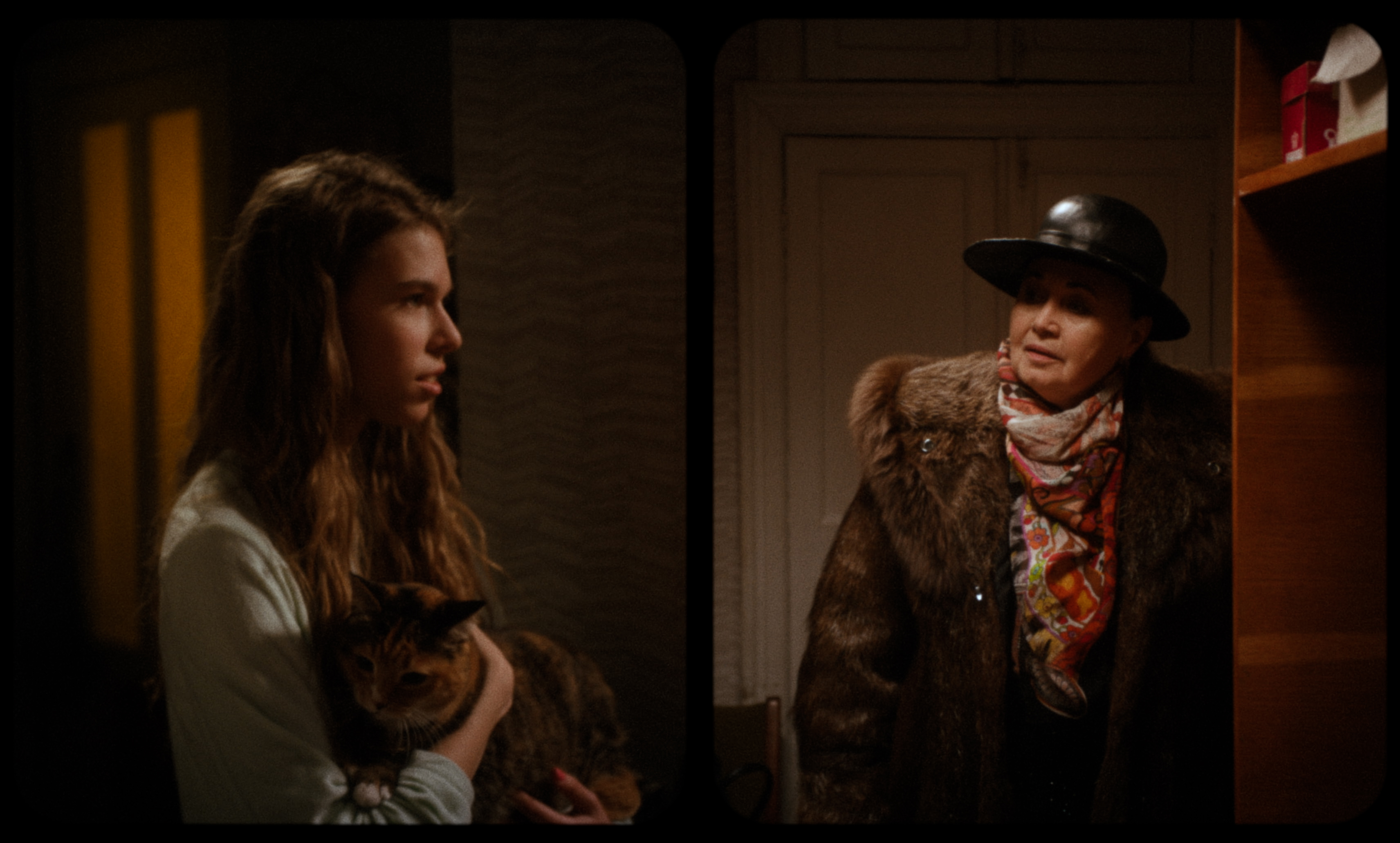 A young woman holding a cat speaks to an older woman in a fur coat and hat at a doorway — a still fr